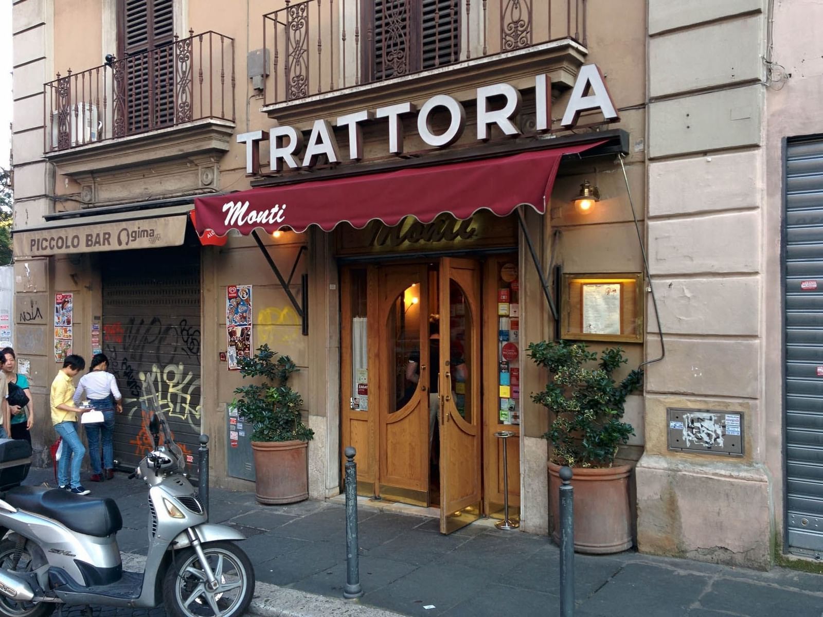 Charming Monti trattoria with a red awning, wooden doors, and potted greenery on a bustling street near The Guardian Hotel