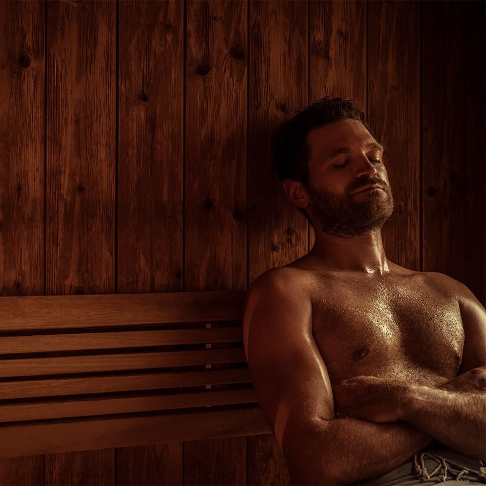 Man with eyes closed, arms crossed, sitting on a bench in a wooden sauna.