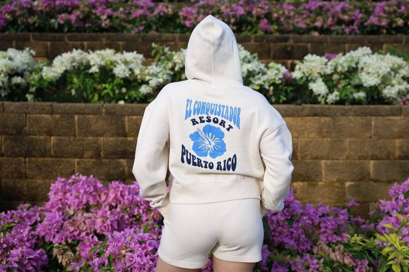 Rear view of a person wearing a white hoodie featuring a blue hibiscus resort logo in front of El Conquistador Resort