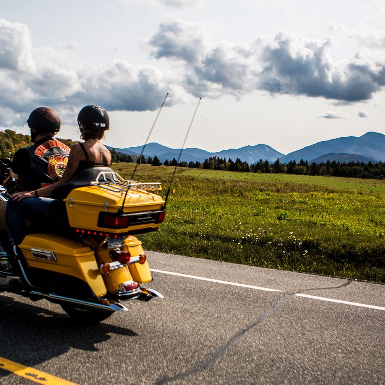 Two people on a yellow motorcycle driving down a road with mountains and green fields in the background.