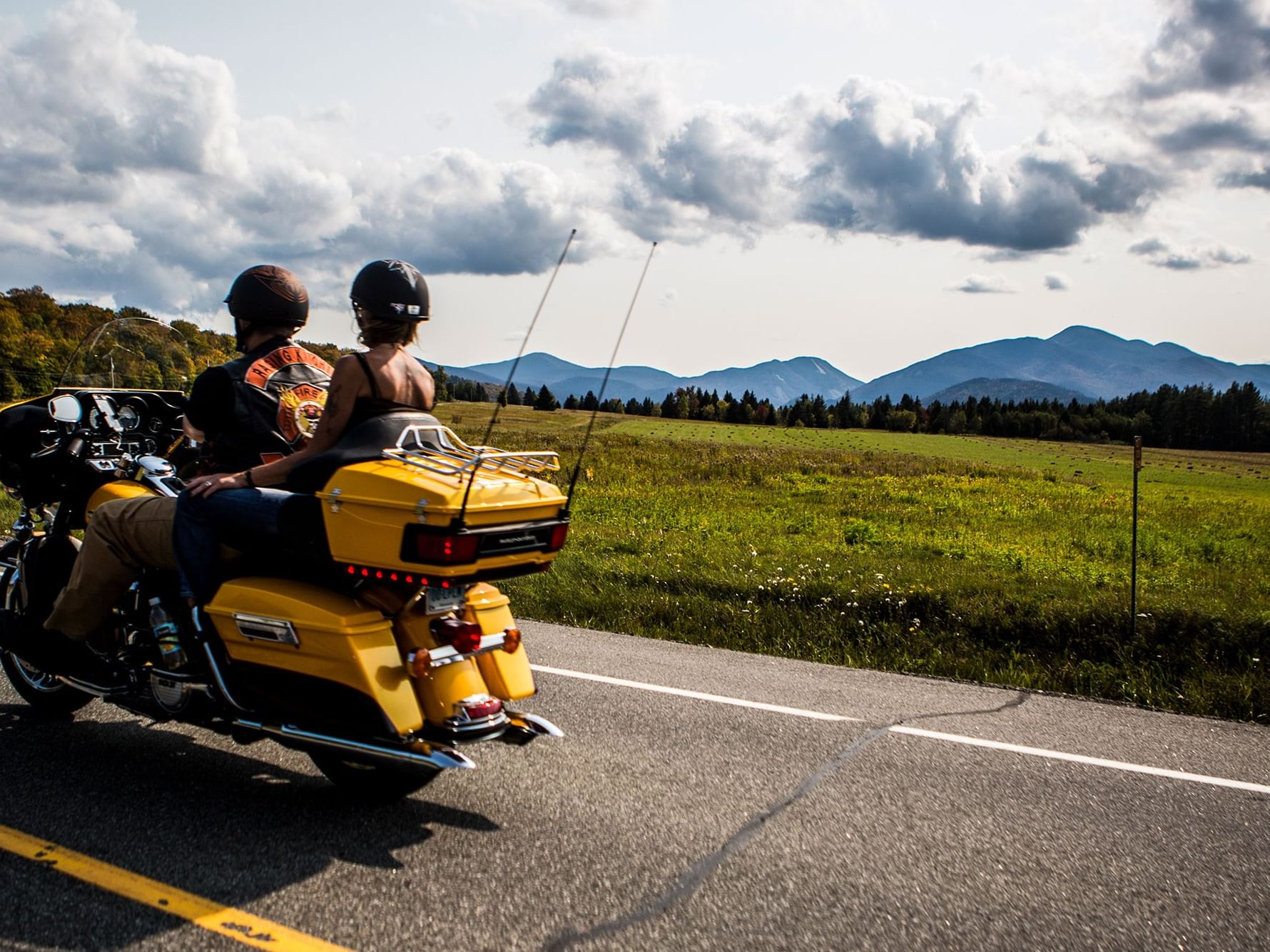Two people on a motorcycle driving down a road with mountains and green fields in the background near High Peaks Resort