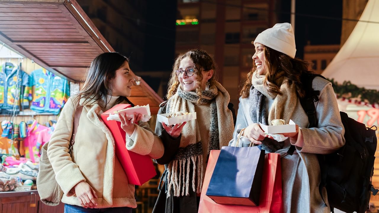 Three women enjoying food and shopping with colorful bags at a Winter Festival & Market