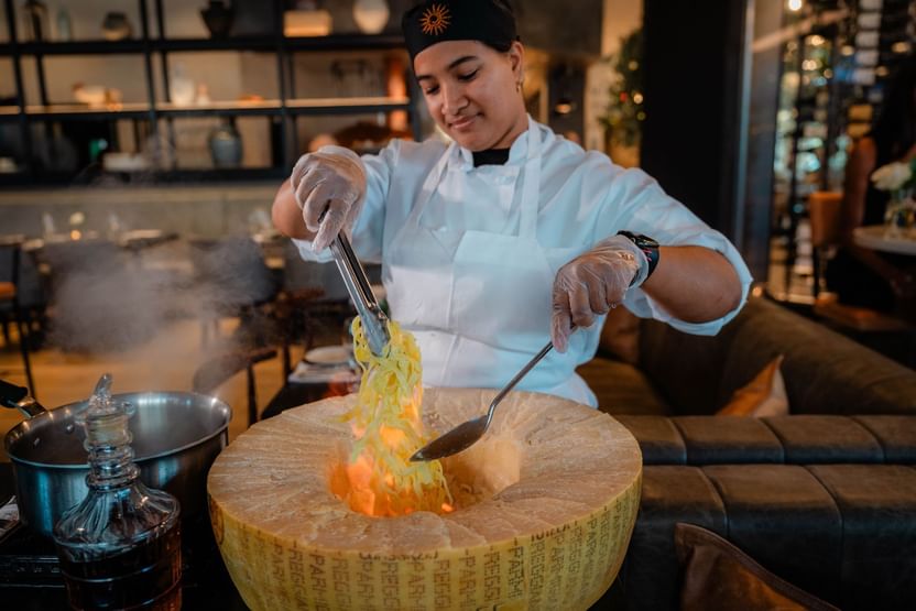 Chef in white uniform and gloves cooking pasta on a giant cheese wheel with flames.