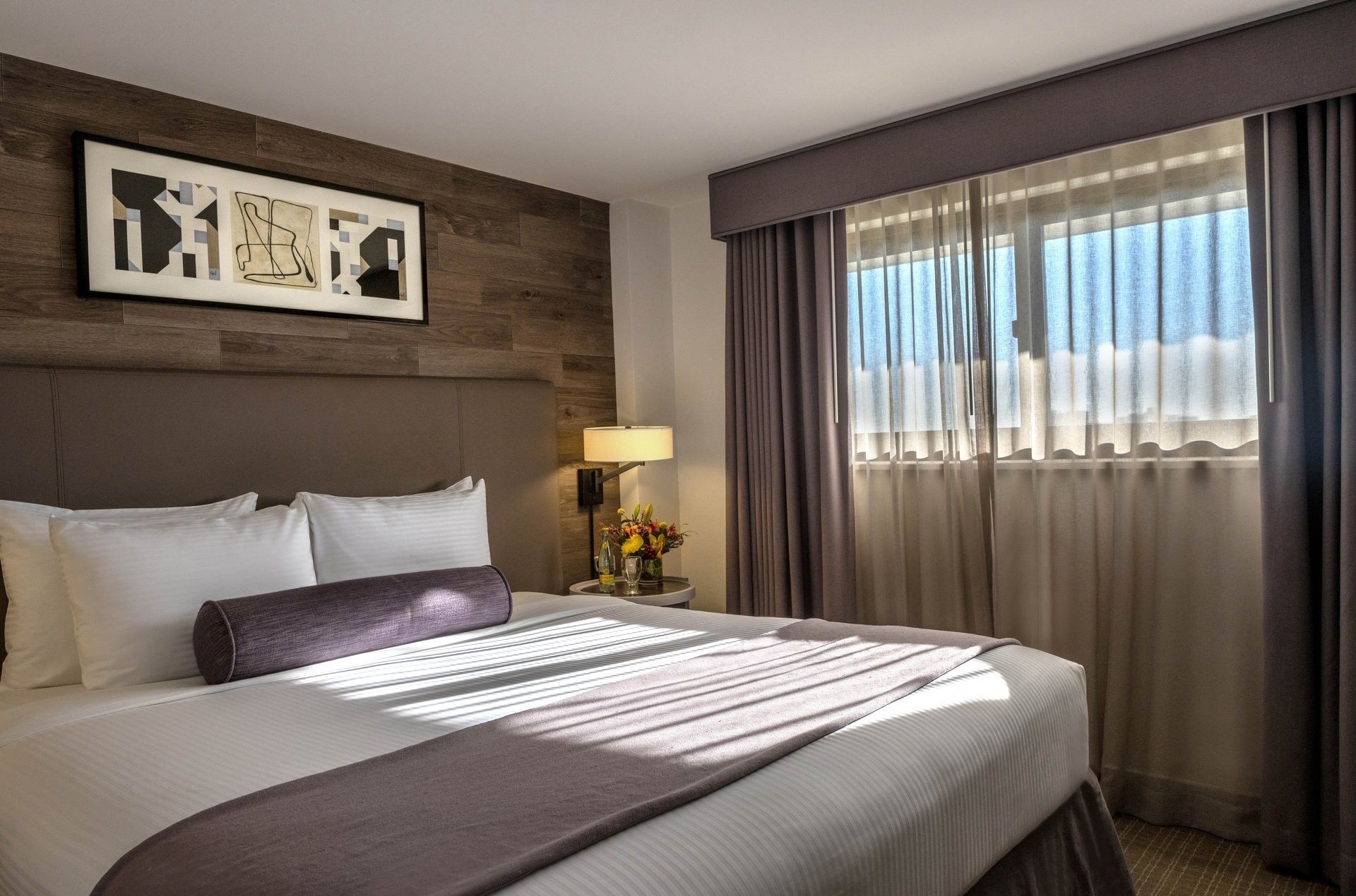 White bed with purple bolster under art on a wood wall by a sunlit window in Premier Suite at Warwick Denver
