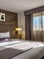 White bed with purple bolster under art on a wood wall by a sunlit window in Premier Suite at Warwick Denver