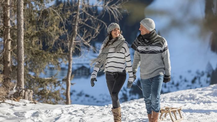 Coppia sorridente in inverno con slittino, neve e alberi sullo sfondo.