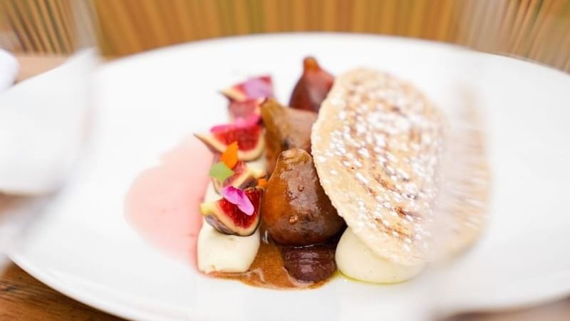 Plate of figs with white sauce, pink sauce, and a powdered pastry piece on a wooden table.