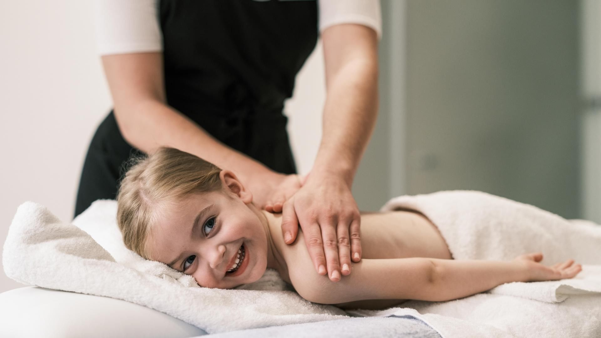 Child lying on a towel, smiling, receiving a shoulder massage from a professional therapist.