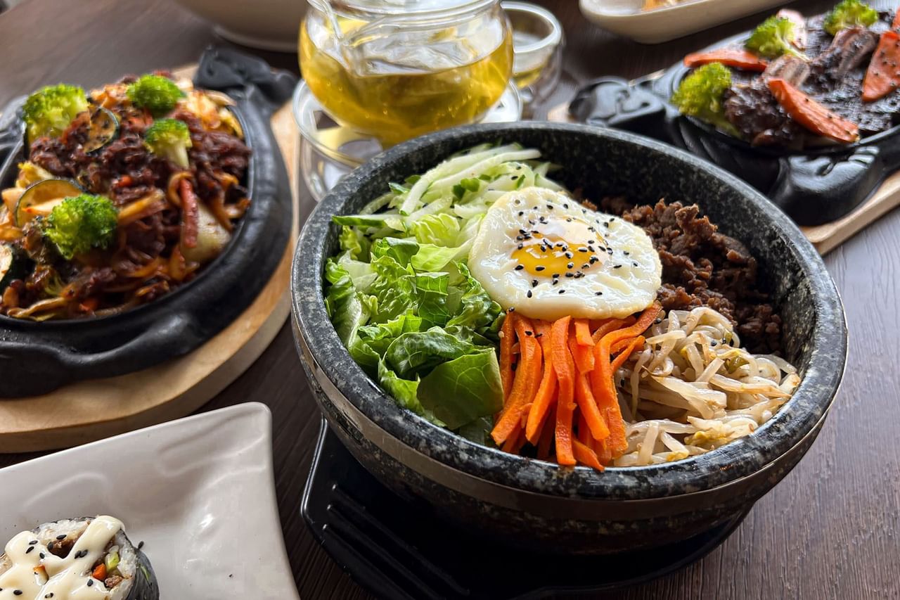 Bowl of Korean bibimbap with egg, veggies, and beef, surrounded by other dishes and a glass of tea.