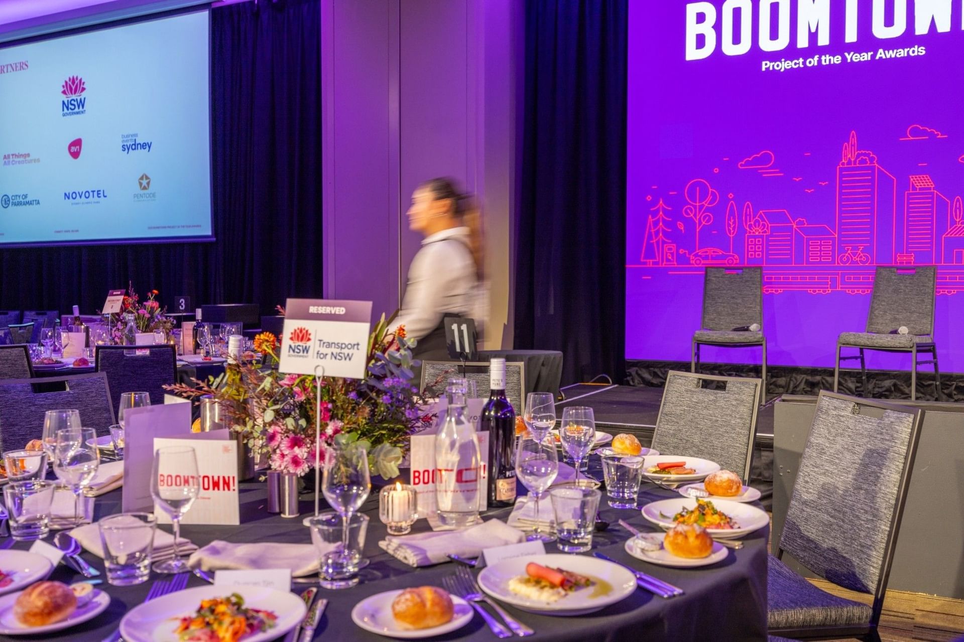 Set a banquet table with a floral centerpiece and wine near the stage in the Freshwater Room at Novotel Sydney Olympic Park