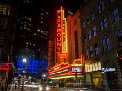 Nighttime urban scene with illuminated Paramount Theatre and surrounding buildings, street lights, and blurred traffic.
