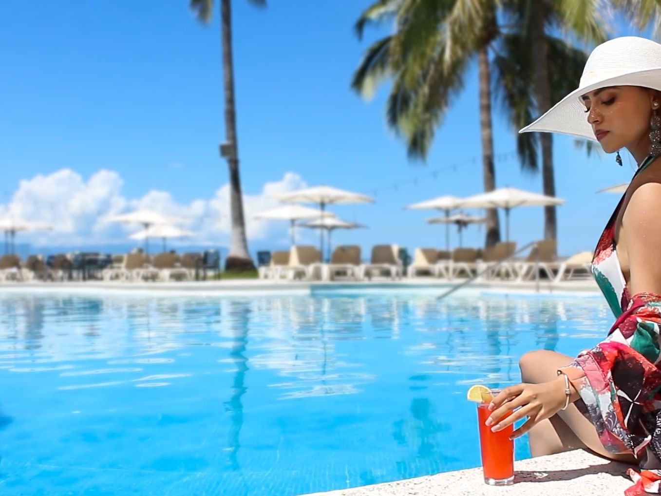 Mujer con sombrero junto a la piscina alcanzando una bebida, con palmeras y tumbonas en Guadalajara Plaza Hoteles & Suites