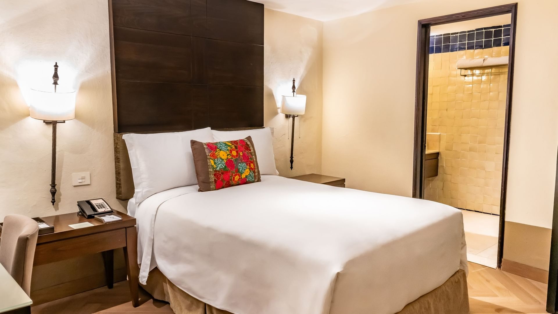 Novicia Matrimonial con cama king, cabecera de madera oscura y cojín bordado colorido en Quinta Real Puebla.
