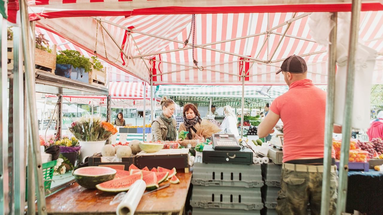 A man in orange shirt stands at a market stall with watermelons and other produce near Coast Lonsdale Quay Hotel 