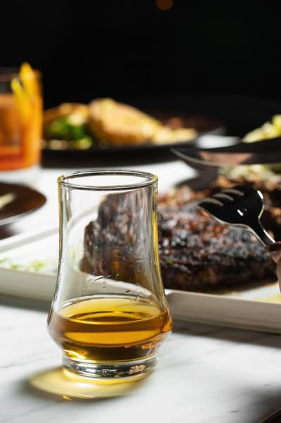 Close-up of a whiskey glass with a fork poised over a charred steak in the background at Cypress & Oak Steakhouse, Frankfort