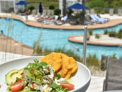 Ceviche dish with avocado and fried plantains served poolside with blue umbrellas and lounge chairs in background