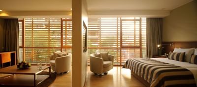 Junior Suite con cama a rayas, sillones y ventanas de madera con vista a áreas verdes en NOI Vitacura