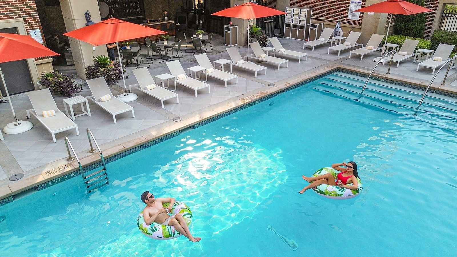 Two guests on leaf-patterned floats by a ladder in a bright blue swimming pool at Warwick Melrose Dallas
