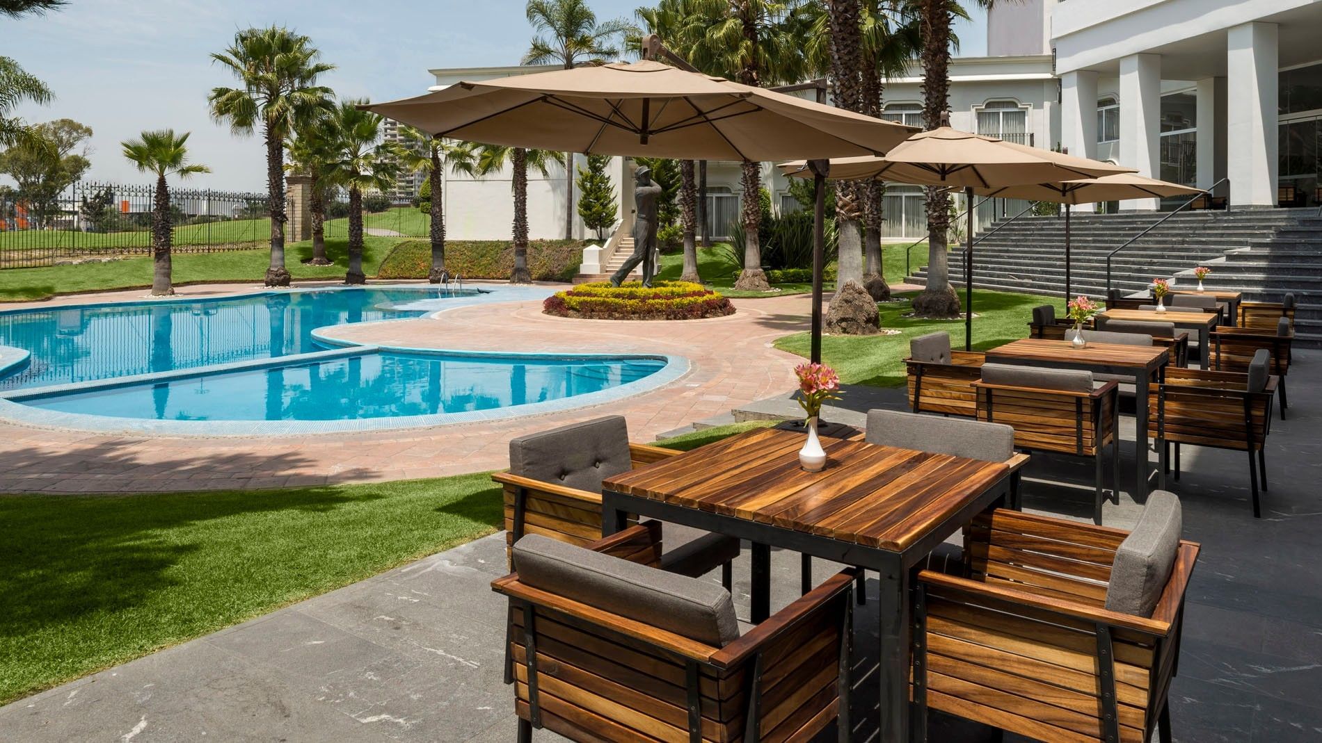 Piscina exterior soleada con mesas de madera y vistas azules cerca de Camino Real Puebla Angelopolis