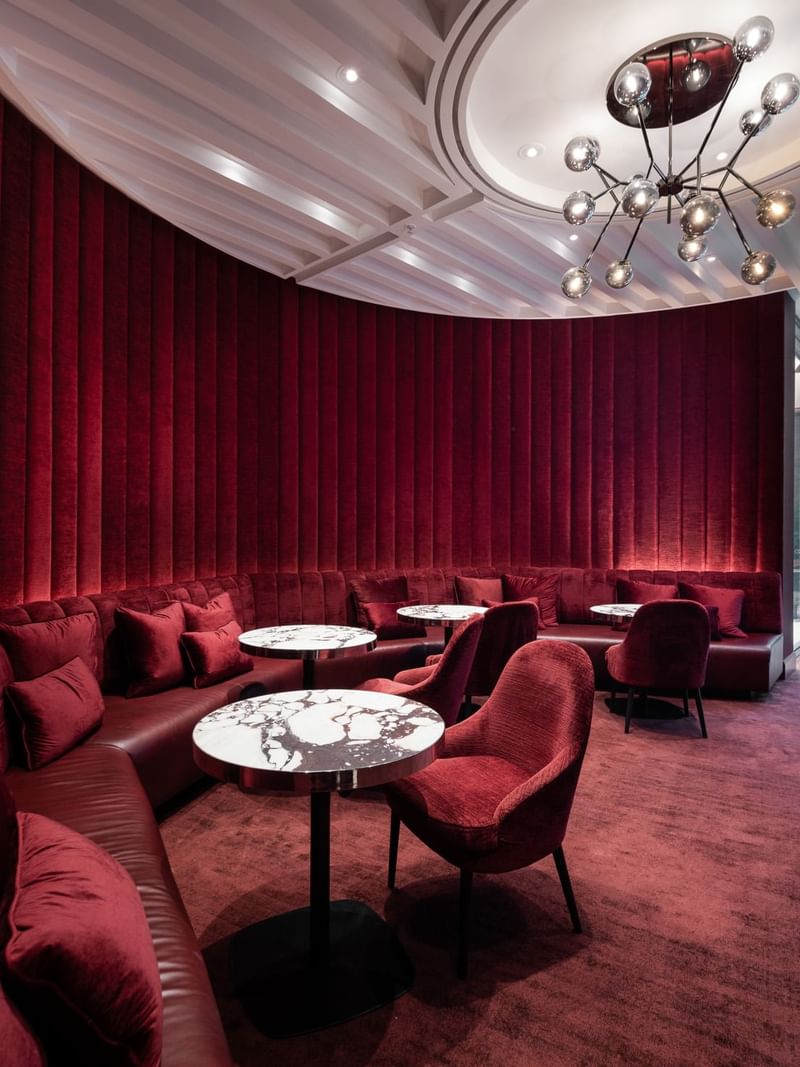 Modern lounge with red cushioned seating, round marble tables, and elegant chandelier, offering a cozy and stylish atmosphere