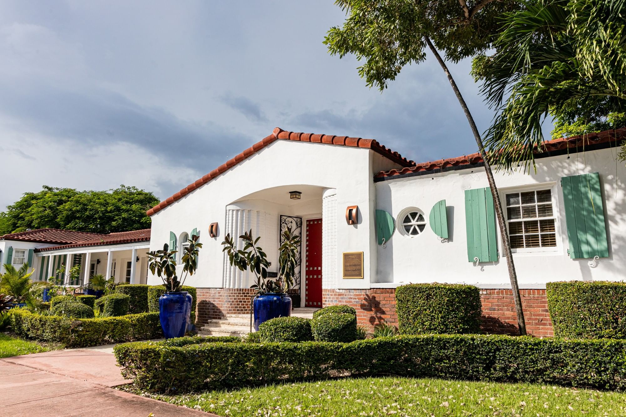 A white stucco building with a red-tiled roof, green shutters, and a red door.