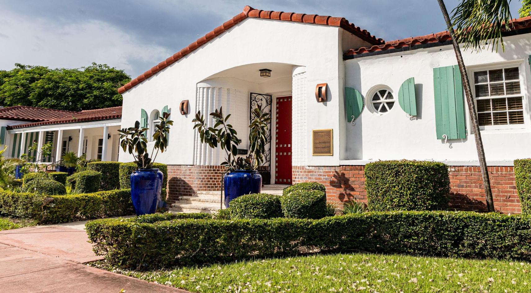 A white stucco building with a red tile roof, green shutters, and a red door, surrounded by manicured hedges and plants.