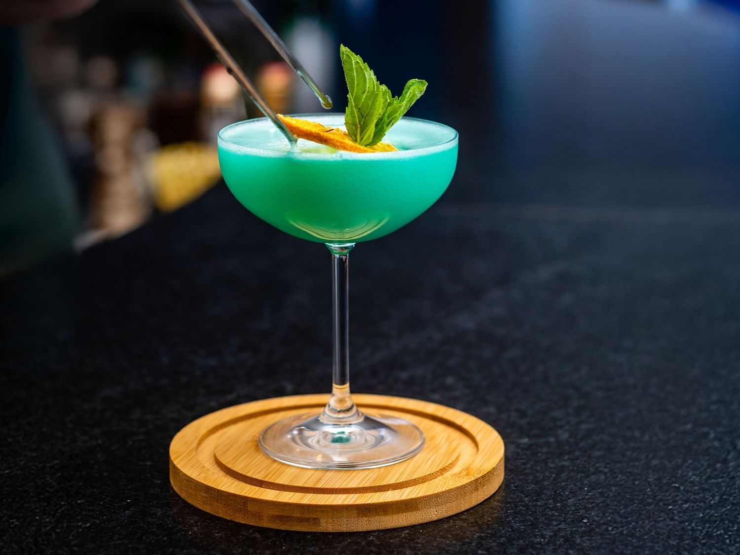 Vibrant blue cocktail in a coupe glass garnished with mint leaves and an orange twist, set on a wooden coaster at Camino Real
