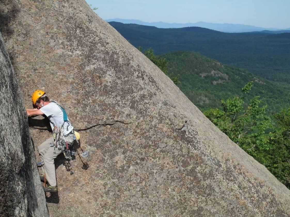 Man in helmet and climbing gear scaling a rock face with forested mountains in background.