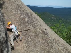 Man in helmet and climbing gear scaling a rock face with forested mountains in background.
