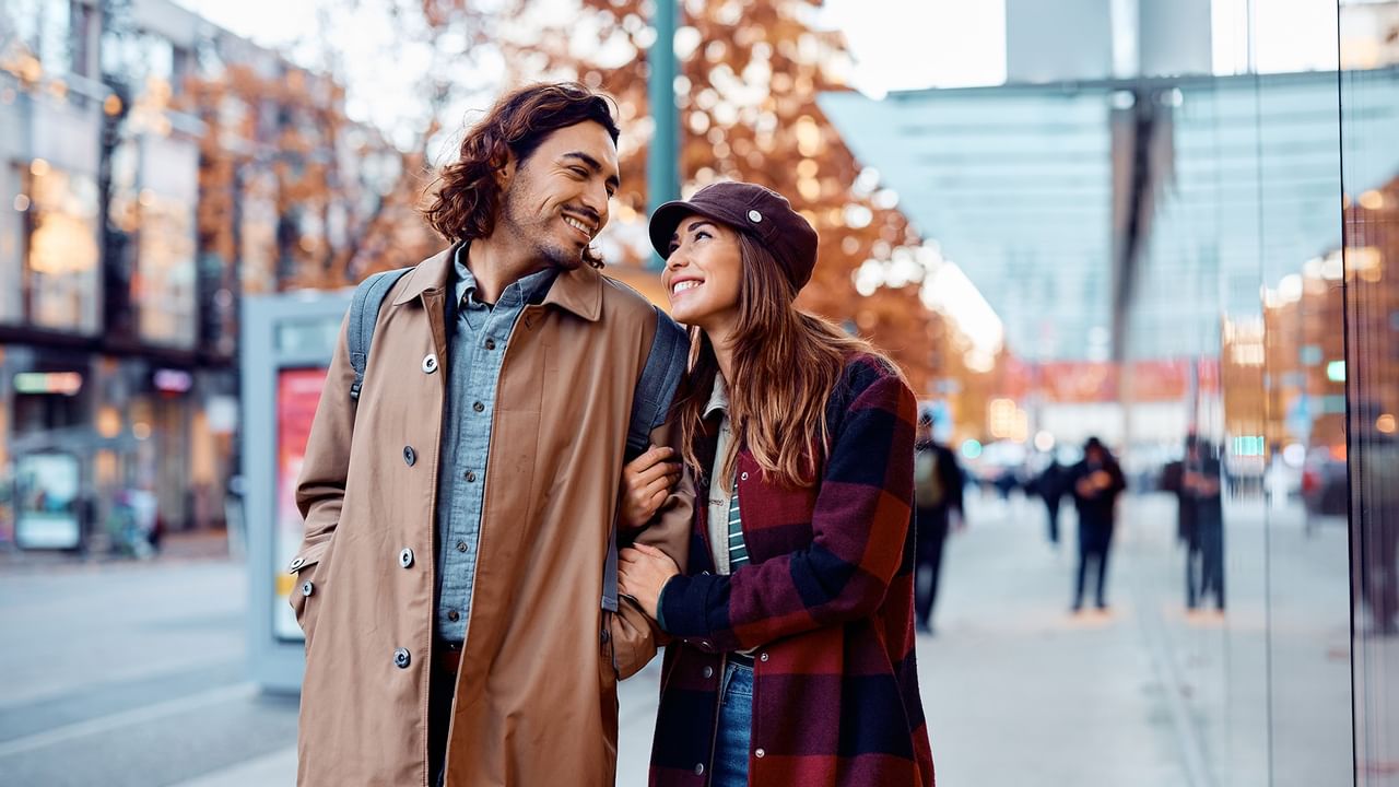 Couple walking arm in arm on a city sidewalk, smiling and looking at each other.