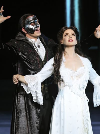A man and woman in Phantom of the Opera costumes on stage with raised arms.