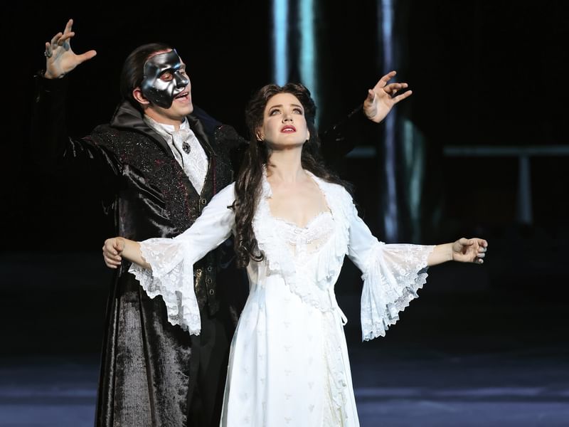 A man and woman in Phantom of the Opera costumes on stage with raised arms.