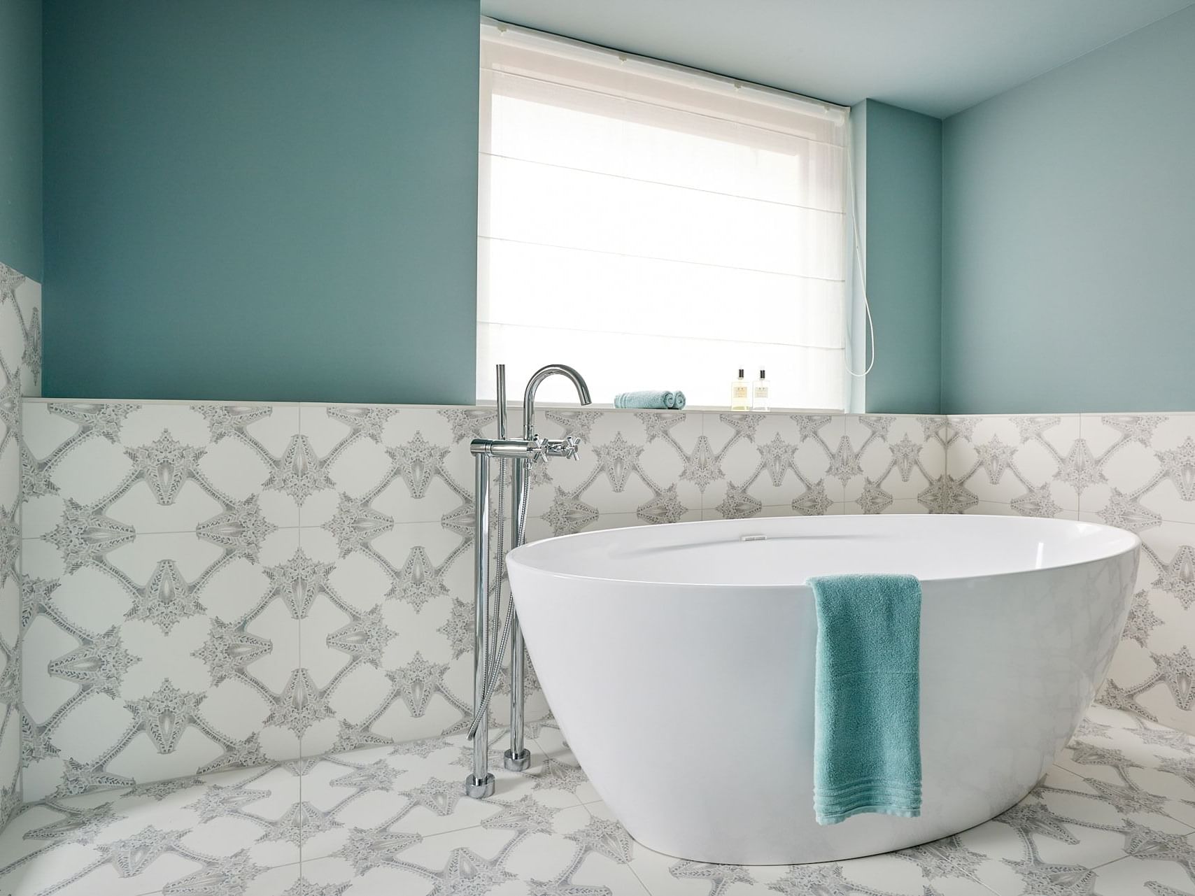 A bathroom with a white freestanding bathtub, blue towel, star-patterned tiles, and a window with blinds.