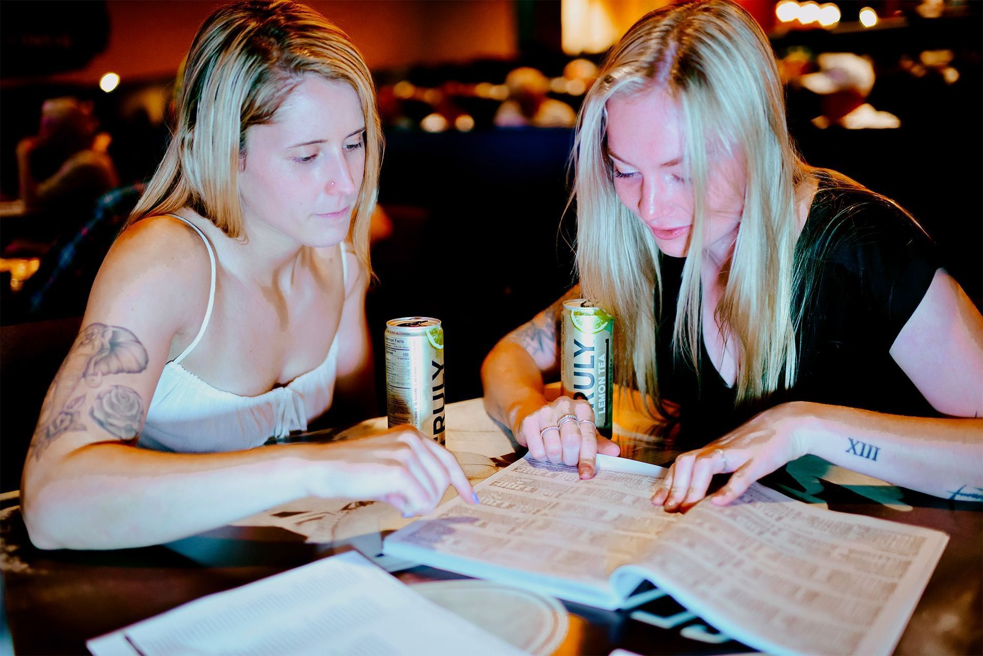 Two guests review a racing form by canned drinks on a table under colorful casino lounge lighting at The Brook