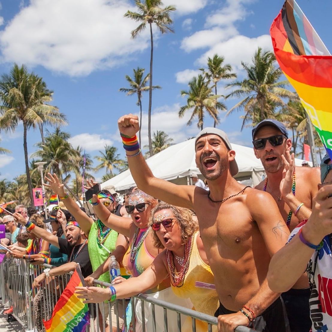 Jubilant crowd celebrating at an outdoor Pride parade with palm trees waving rainbow flags near Tradewinds Apartment Hotel