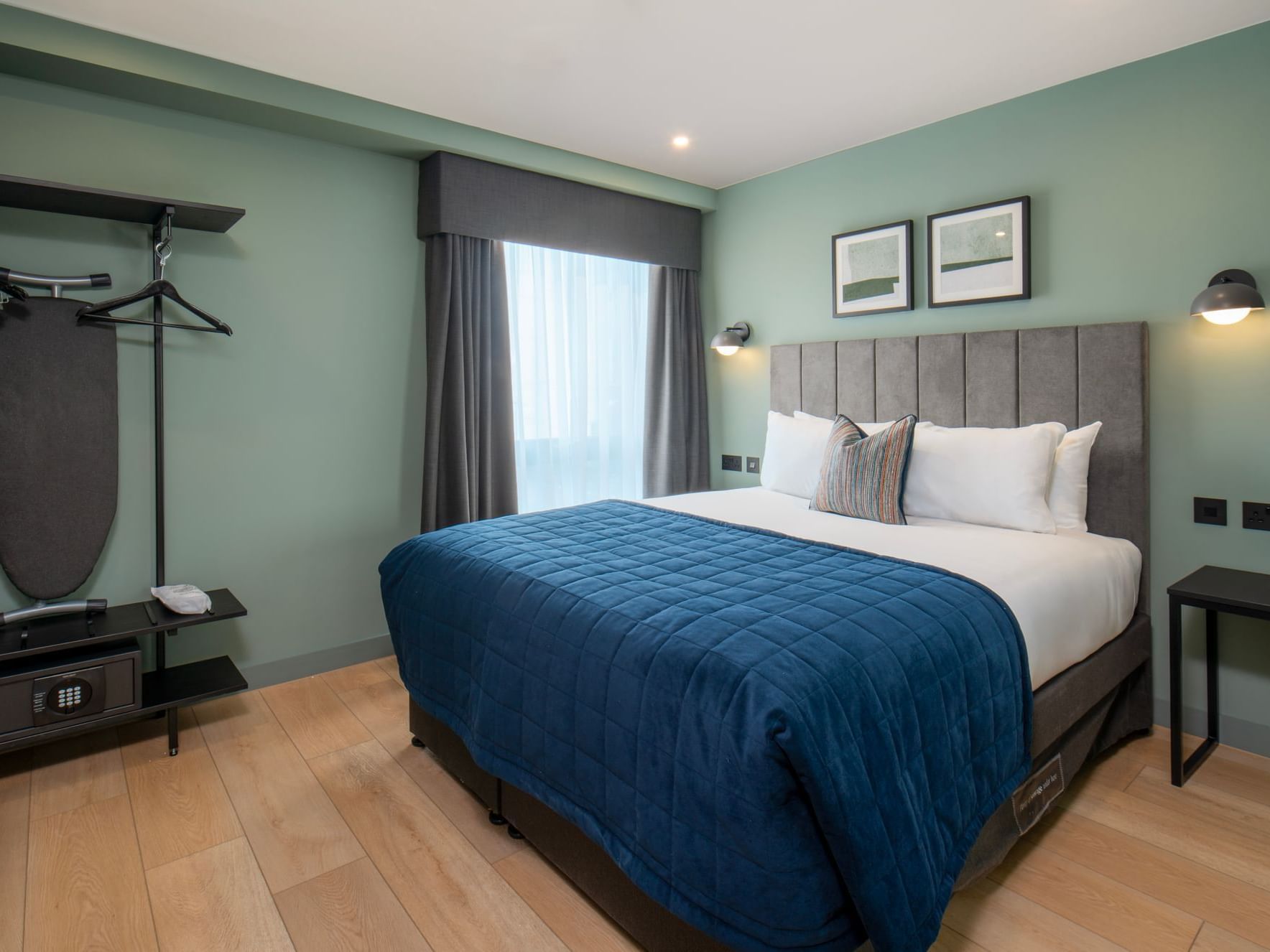 Bedroom with side tables, open wardrobe with safe, large windows, natural light green walls and blue quilted throw