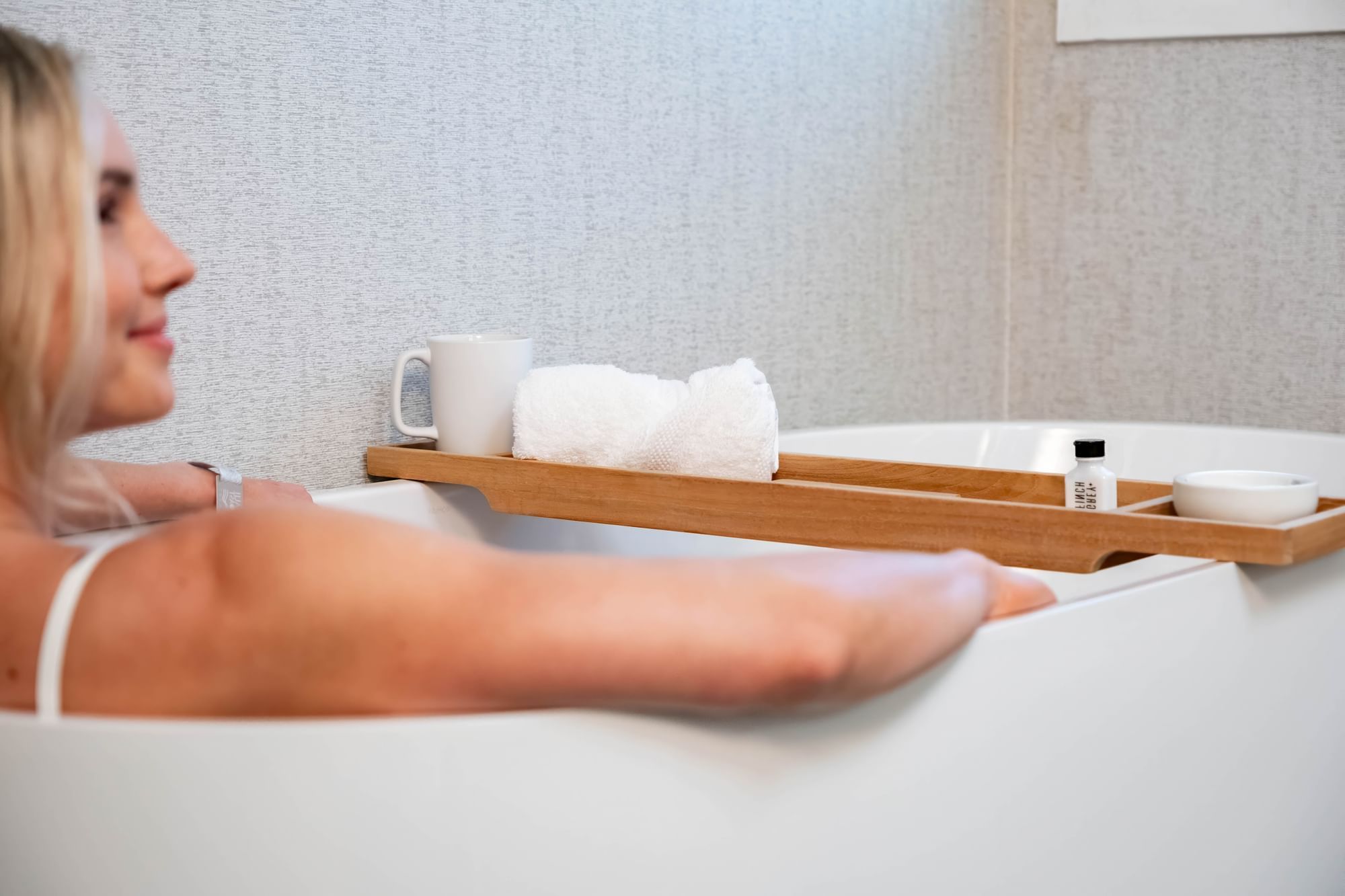 a woman relaxing in a bathtub with a wooden tray holding a mug, towels, and bottles