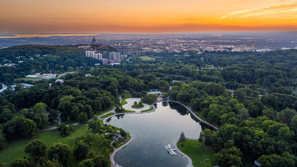 Vue aérienne d'un parc montréalais avec un lac et la silhouette de la ville au coucher du soleil.