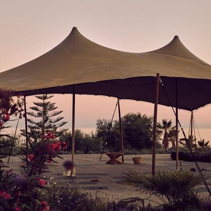 Large tent with peaked tops and wooden supports set on a deck with plants and trees.