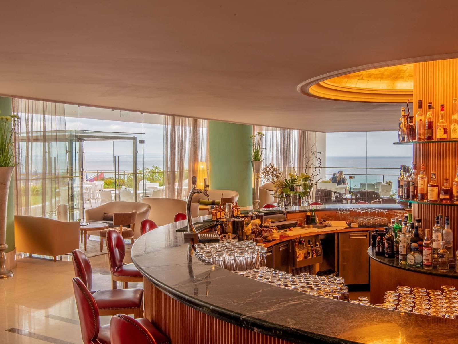 Bar circulaire élégant avec vue sur l'océan, nombreux fauteuils et chaises, et étagères bien garnies de bouteilles.