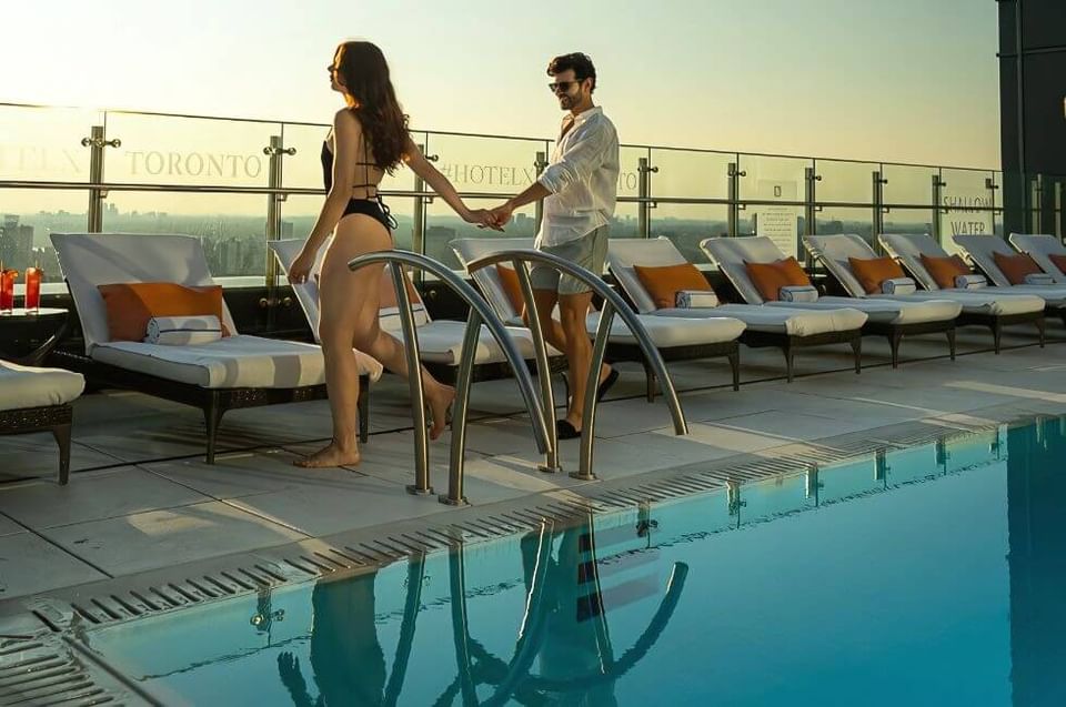 Couple walking by a rooftop pool with cushioned loungers under warm sunset light on the pool terrace at Hotel X Toronto