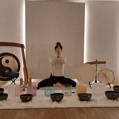 Woman in meditation pose with hands in prayer position surrounded by bowls and gongs on a rug.