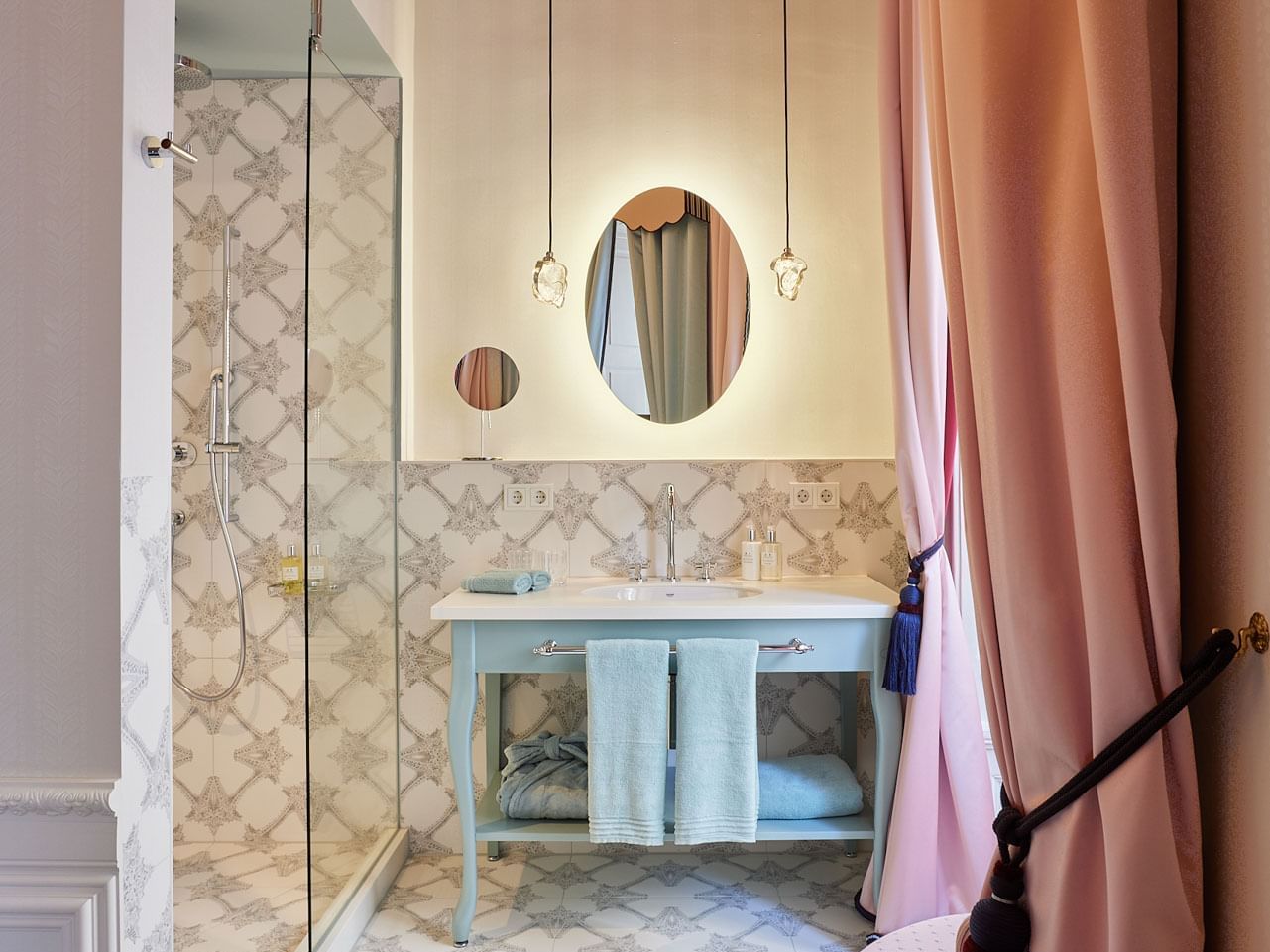 A bathroom with a shower, mirror, blue vanity, patterned tiles, and pink curtains.
