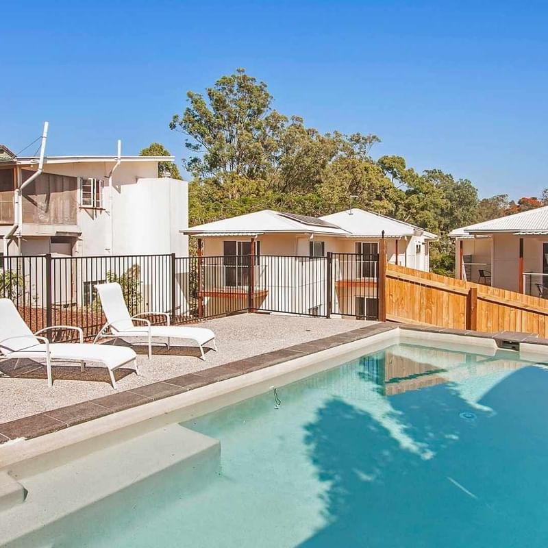 Outdoor swimming pool with sun loungers surrounded by residential buildings and trees under a clear blue sky.