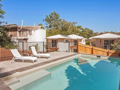 Outdoor swimming pool with sun loungers surrounded by residential buildings and trees under a clear blue sky.
