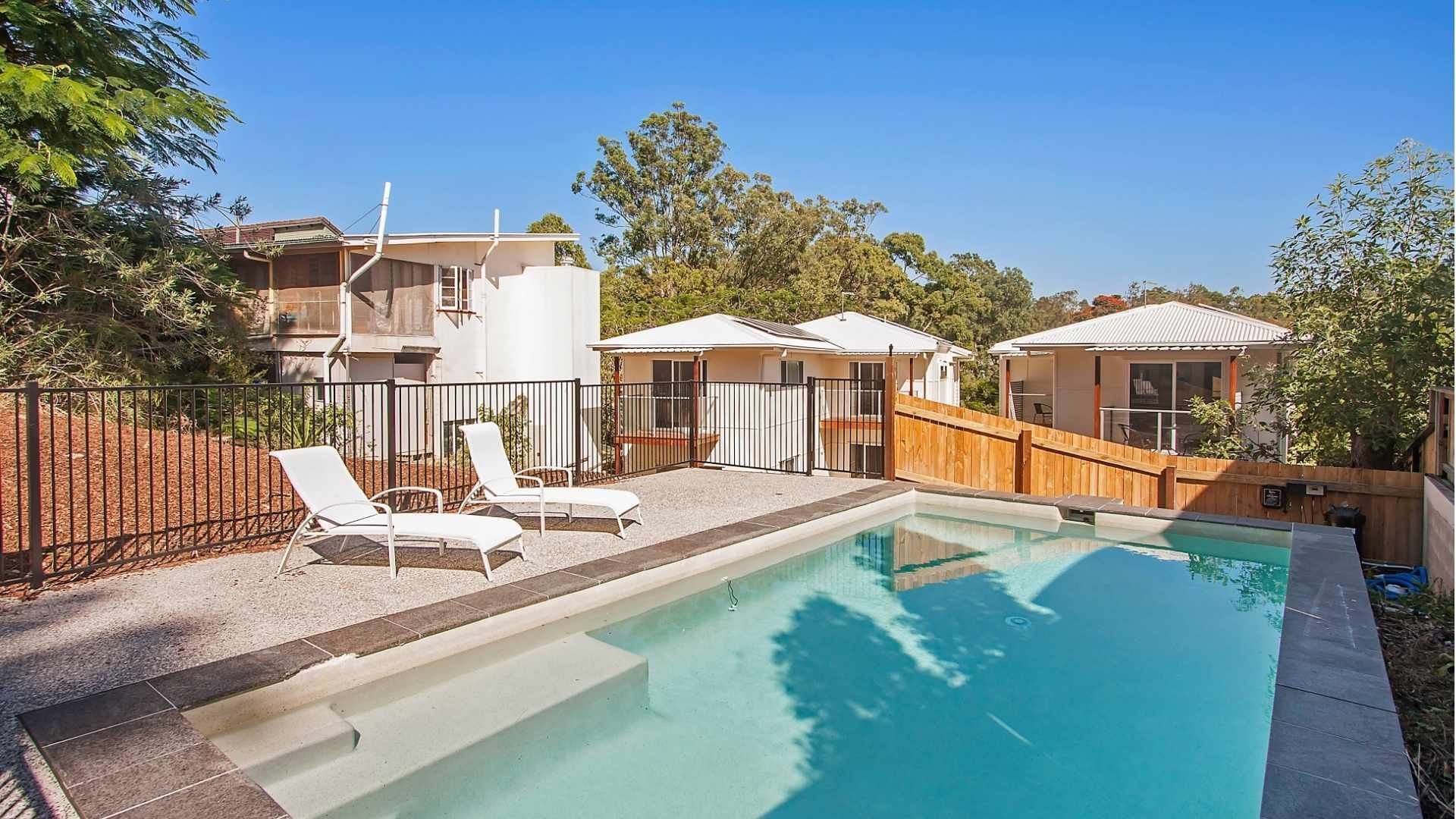Outdoor swimming pool with sun loungers surrounded by residential buildings and trees under a clear blue sky.