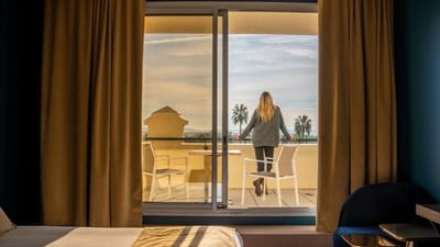 Woman with blonde hair standing on balcony looking out at ocean through sliding glass doors.