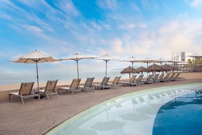 Piscina con tumbonas, sombrillas y gazebos de techo de palma en el Sunset Plaza Beach Resort durante un atardecer con vistas a la playa de Puerto Vallarta