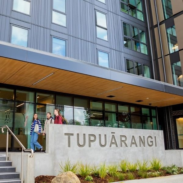 Modern building entrance with concrete Tupuārangi sign, glass doors, and students walking on steps and pathway