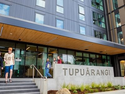 Modern building entrance with concrete Tupuārangi sign, glass doors, and students walking on steps and pathway.